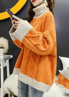 a woman standing in front of a wall holding a cell phone and wearing an orange sweater