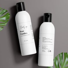 two bottles of body lotion next to a green plant on a gray surface with black caps