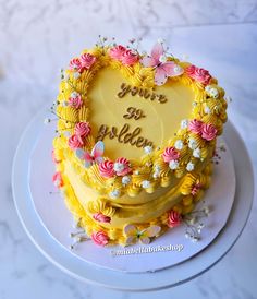 a heart shaped yellow cake with pink and white flowers on it's side, sitting on a plate