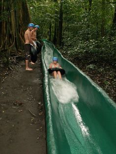 two men are riding down a water slide in the woods, one is on his knees