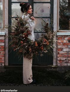 a woman standing in front of a brick building holding a wreath with pine cones on it