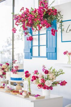 a table topped with cakes and flowers next to blue shutters on the wall behind it