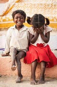 two young children sitting on a ledge with their hands covering their faces and smiling at the camera