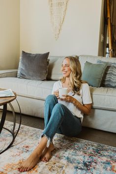 a woman sitting on the floor in front of a couch holding a coffee cup and looking at her phone