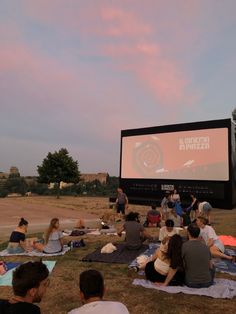 a group of people sitting on top of a field next to a large projection screen