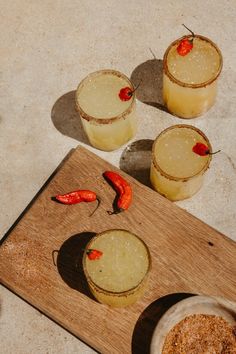 three glasses filled with drinks sitting on top of a wooden cutting board next to chili peppers