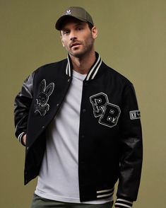 A man models the MENS ROBBINS VARSITY BOMBER JACKET - B6J524Z1OW by Psycho Bunny, featuring white accents and chenille patches like a rabbit skull and crossbones. He complements the look with a green cap and white t-shirt, all set against a muted green backdrop. Black Varsity Jacket Outfit Men, Varsity Jacket Outfit Mens, Varsity Fashion, Black Varsity Jacket, Varsity Jacket Outfit, Jacket Sleeves, Streetwear Outfit Ideas, Leather Varsity Jackets, Jordan Outfit