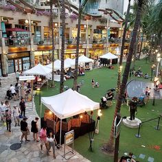 many people are walking around in an open area with tables and umbrellas on the grass