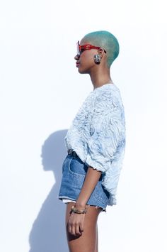 a woman with green hair and sunglasses standing in front of a white wall wearing denim shorts