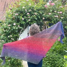 a woman is holding up a crocheted shawl in front of some flowers