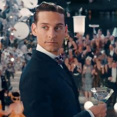 a man in a tuxedo holding a martini glass with an audience behind him