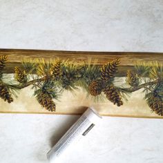 a piece of wood with pine cones and branches painted on it, next to a tube of toothpaste