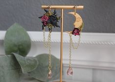 a pair of earrings with flowers and chains hanging from them on a stand next to a potted plant