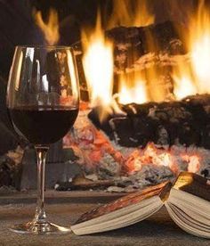 a glass of wine and an open book sitting in front of a fireplace with flames