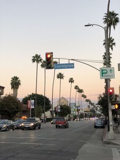 cars are driving down the street in front of palm trees and buildings at dusk with red traffic lights