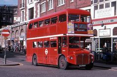 a red double decker bus driving down a street next to tall buildings and people on the sidewalk