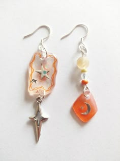 two pairs of earrings with charms attached to them on a white surface, one has a star and the other has a moon