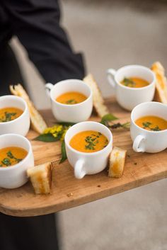 a wooden tray topped with cups of soup next to toasted bread on top of a table