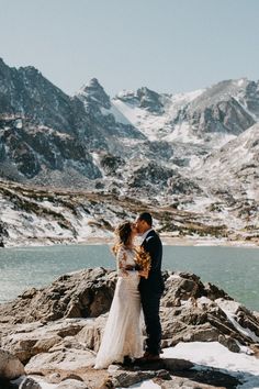 a bride and groom standing on rocks in front of snow covered mountains with their arms around each other