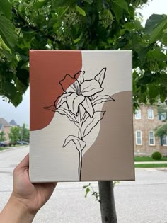 a hand holding up a piece of art that has flowers on it and is in front of a tree