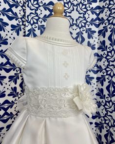 A traditional off-white dress with pleats and little roses embroidered on the front of the body, the sash has gorgeous lace on the sash. The skirt has beautiful details throughout. The flower is an applique, which can be removed at any time. It has covered buttons on the back for closure and a bow. Made in Spain Dry Clean 100% silk Final sale, no exchanges nor returns are available Elegant Fitted Dress For Confirmation, Elegant Fitted Confirmation Dress, Fitted Lace Bodice Dress For Confirmation, Fitted Floral Embroidery Baptism Dresses, Fitted Floral Applique Dress For Confirmation, Elegant Floral Applique Dress For Baptism, White Satin Confirmation Dress, Formal Lace Dress With Pleated Bodice, White Satin Dress With Pleated Bodice