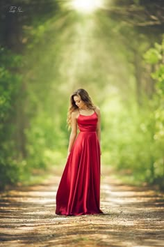 a woman in a long red dress standing on a dirt road surrounded by green trees