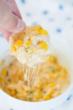 a hand holding a tortilla chip over a bowl of corn and cheese dip