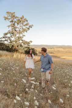 a man and woman holding hands walking through tall grass in the middle of a field