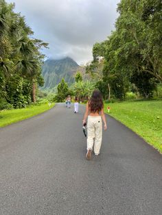 a woman walking down the middle of a road next to lush green trees and mountains