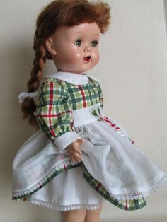 a doll with red hair wearing a white dress and green plaid shirt is standing on a white surface