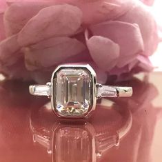 an emerald and diamond ring sitting on top of a table with pink flowers in the background