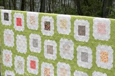 a green and white quilt hanging from a wooden fence with trees in the back ground