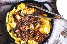 a skillet filled with pasta and meat on top of a wooden table next to utensils