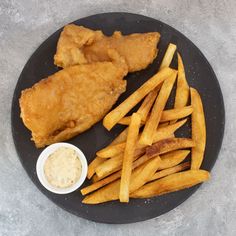 fried fish and french fries on a black plate with dipping sauce next to it,