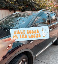 a person holding up a sticker that says silly goose on the loose in front of a car