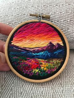 someone is holding up a small embroidery art piece with mountains in the background and colorful flowers on it