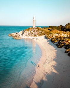 two people are walking on the beach towards a light house in the distance with blue water