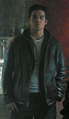 a man standing in front of a brick wall wearing a black leather jacket and white t - shirt