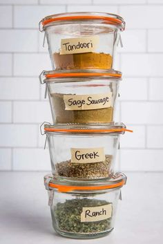 three glass containers filled with spices and labeled in different labels on the lids are stacked up against a white brick wall