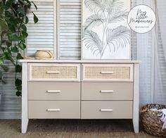 a white dresser with wicker drawers next to a plant and a painting on the wall