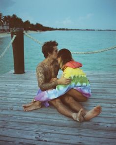 a man and woman sitting on a dock next to each other with their arms around each other