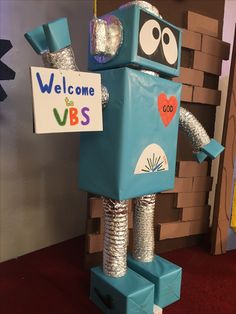 a cardboard robot holding a welcome sign in front of a brick wall