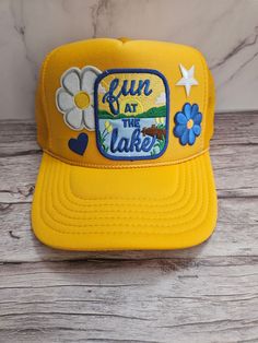 Lake Day Patch Trucker Hat The front of this hat is embellished with patches including one that says fun at the lake, flowers and a heart. This hat is perfect for a day out at the lake or day on the boat. The blue and yellow colors are perfect for summer! The hat has a foam front panel and a mesh back with and adjustable strap in the back. Retro Summer Hats For Outdoor Activities, Fun Outdoor Hat For Spring, Fun Outdoor Spring Hat, Yellow Outdoor Hat For Spring, Fun Spring Hats For Outdoor Activities, Fun Spring Outdoor Hats, Yellow Spring Outdoor Hat, Fun Summer Trucker Hat With Embroidered Patch, Yellow Trucker Hat Baseball Cap For Spring