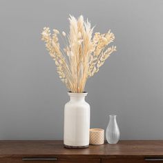 a white vase sitting on top of a wooden table next to a vase filled with dry grass