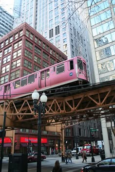 a pink train traveling over a bridge in the middle of a busy city with tall buildings