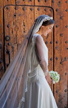 a woman in a wedding dress and veil standing next to a wooden door holding a bouquet