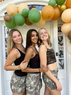 three young women are posing for the camera in front of a balloon arch with green, yellow and orange balloons