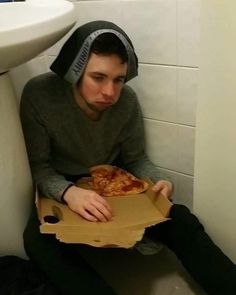 a man sitting on the toilet with a pizza in his hand and wearing a hoodie