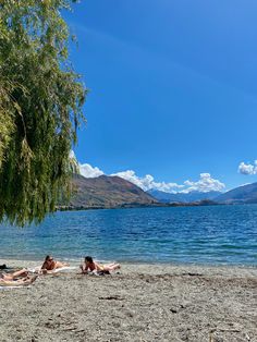 two people laying on the beach under a tree next to some water with mountains in the background