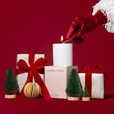 a person lighting a candle surrounded by christmas decorations and gifts on a red background with white feathers